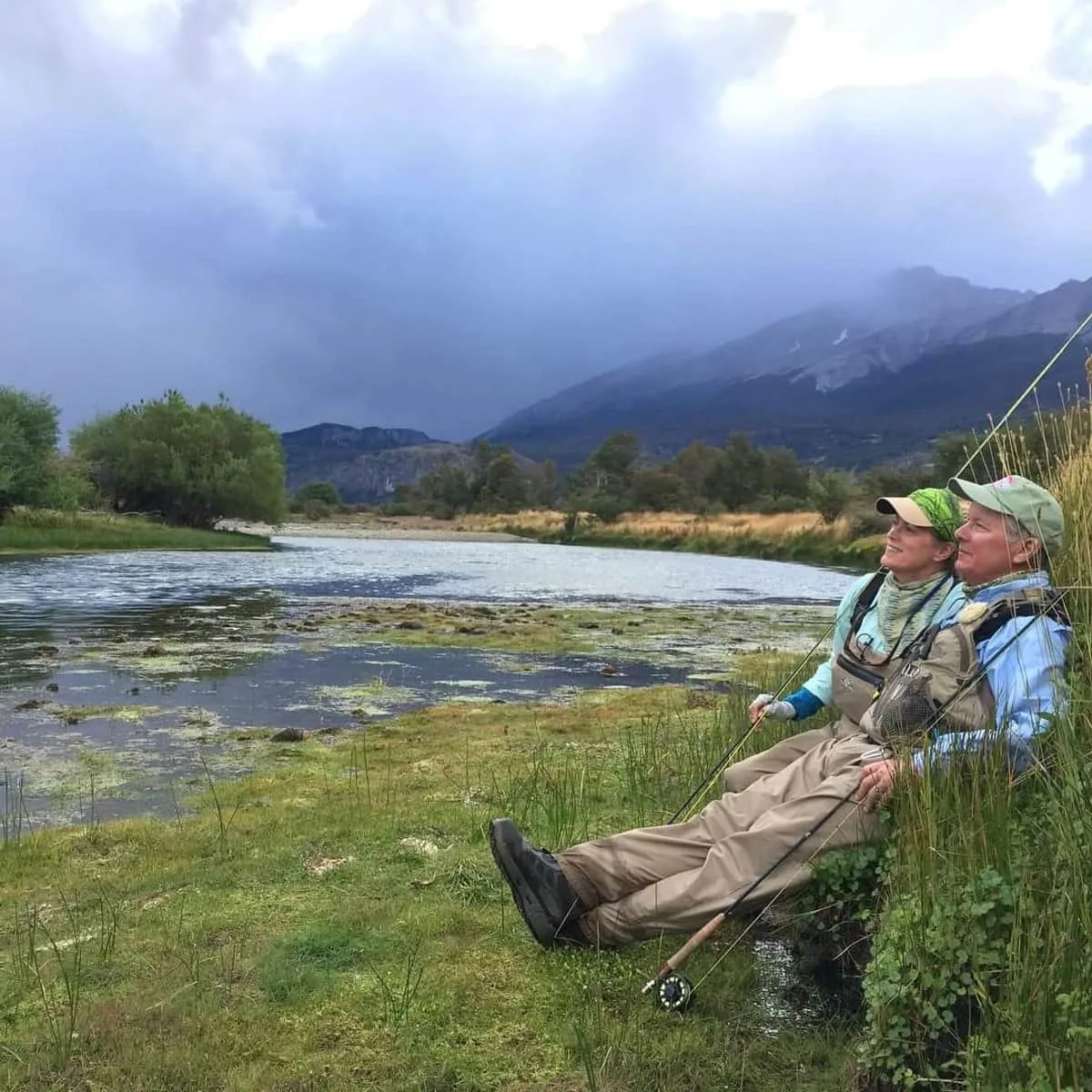 Anglers resting on a grassy riverbank under Patagonia mountains at El Saltamontes Lodge